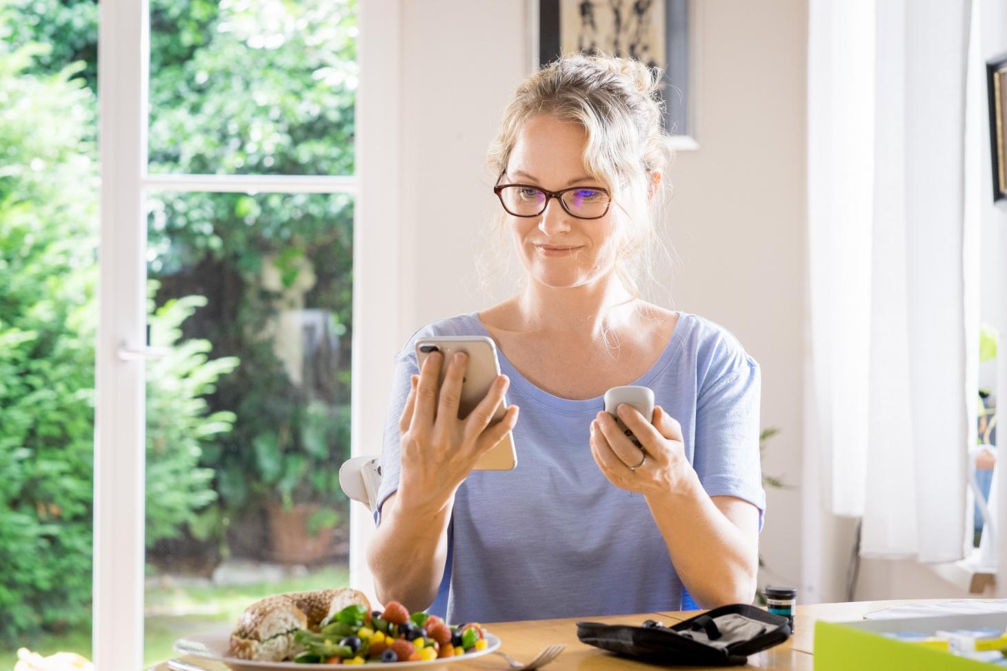 Woman wearing purple shirt and wearing glasses sitting at table next to window. Holding phone and Accu-Chek® Guide meter and there is a plate of food, half a bagel and a salad on the table. There are test strips on the table as well and the corner of the Bundle box.